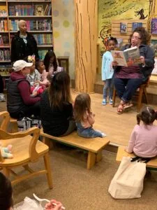 Children and adults gathered in a library during a story time reading session, with bookshelves in background and a woman reading aloud to the group.