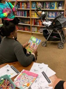 Woman reading picture book at table in library with bookshelves and stroller in background