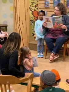 Adult reading picture book to group of children sitting on floor in classroom with animal artwork on wall