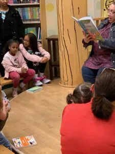 An adult reading a picture book aloud to a group of children seated on the floor in a library with bookshelves visible in the background.