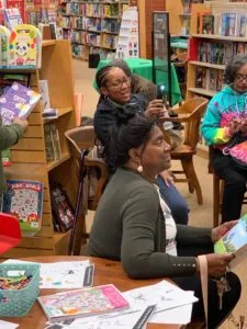 Children and adults seated in a library surrounded by bookshelves, with books and activity materials on the table in front of them.