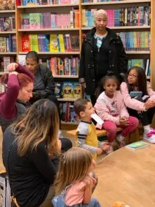 Adult and children gathered in a library with colorful bookshelves in the background during what appears to be a reading or educational event