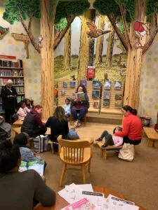 Children and adults seated in a library reading nook decorated with large painted trees, listening to a story or book reading session