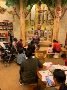 Children and adults gathered in a library reading area with forest-themed wall mural during a storytelling session