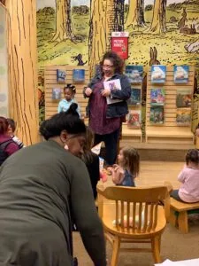 Adult reading picture book to children seated in wooden chairs during library storytime with forest mural background