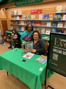 Author at green table holding children's book at bookstore event with shelves of books behind