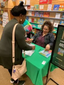 Two women at a green table in a bookstore with shelves of children's books behind them during what appears to be a book signing event.