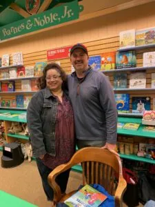 Two people smiling at a bookstore display with shelves of children's books behind them