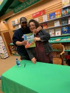 Two people posing together at a green table in a bookstore, holding a children's book titled Bluebird & Friends