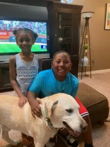 Two children petting a white Labrador Retriever in a living room with a television playing in the background.