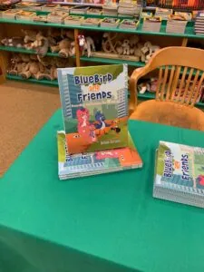 Children's books titled 'Bluebird and Friends' displayed on a green table in a library or classroom setting with shelves of stuffed animals and books visible in the background