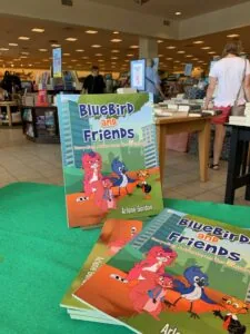 Children's book "BlueBird and Friends" displayed on green table in library with books and patrons in background