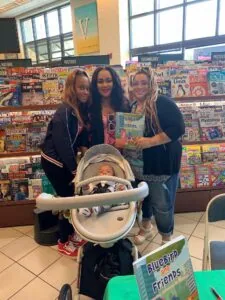 Three women posing together in front of a wall of magazine covers, with an infant in a stroller in the foreground