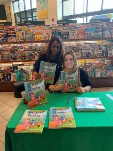 Two women holding Bluebird Friends books at a green table display in a library or educational setting with magazine covers on wall behind them