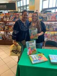 Two women smiling at a table with children's books, displaying 'Blue Bird and Friends' publications in front of a magazine display wall