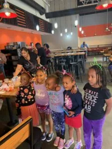 Five young girls wearing decorative headbands stand together at a children's party in a bright orange and white venue with hanging lights.