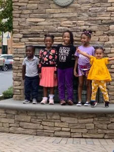 Five young children standing together on a concrete ledge against a stone wall, smiling at the camera