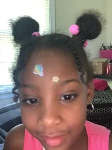 Child with two black puffs styled with pink pom-poms wearing pink top with face stickers