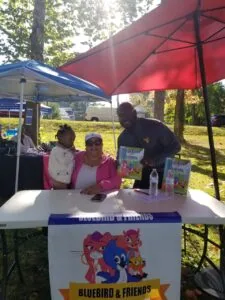 Three people at an outdoor event booth under red and blue umbrellas, with Bluebird & Friends signage and merchandise on display