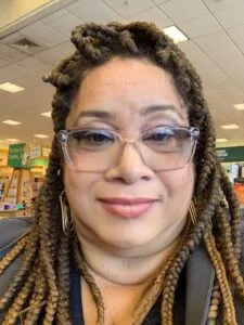 Person wearing clear-framed glasses and gold hoop earrings with braided hair styled in buns, smiling at camera in front of classroom backdrop