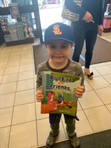 Young child wearing blue baseball cap and camouflage shirt holding Bluebird and Friends book in indoor retail space