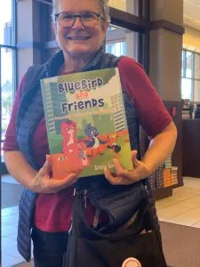 Person wearing glasses and colorful vest holding a children's book titled 'BlueBird and Friends' indoors with windows in background