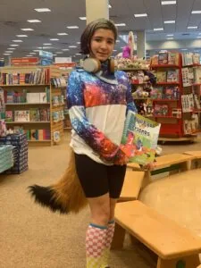 Child wearing galaxy-patterned hoodie and colorful socks holds a children's book in a library with bookshelves behind them