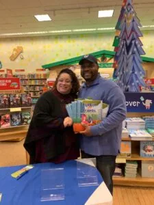 Two people holding a Bluebird Friends book in a library with colorful decorations and bookshelves