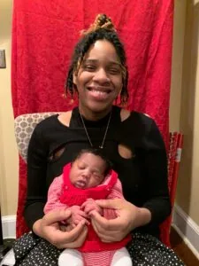 Smiling woman holding sleeping infant in pink outfit against red backdrop