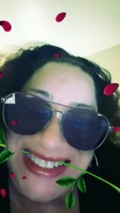 Person wearing dark aviator sunglasses and red lipstick, smiling at camera with green plant stem and red petal decorations