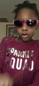 Child wearing pink sunglasses with rhinestone details and burgundy sparkle squad graphic shirt